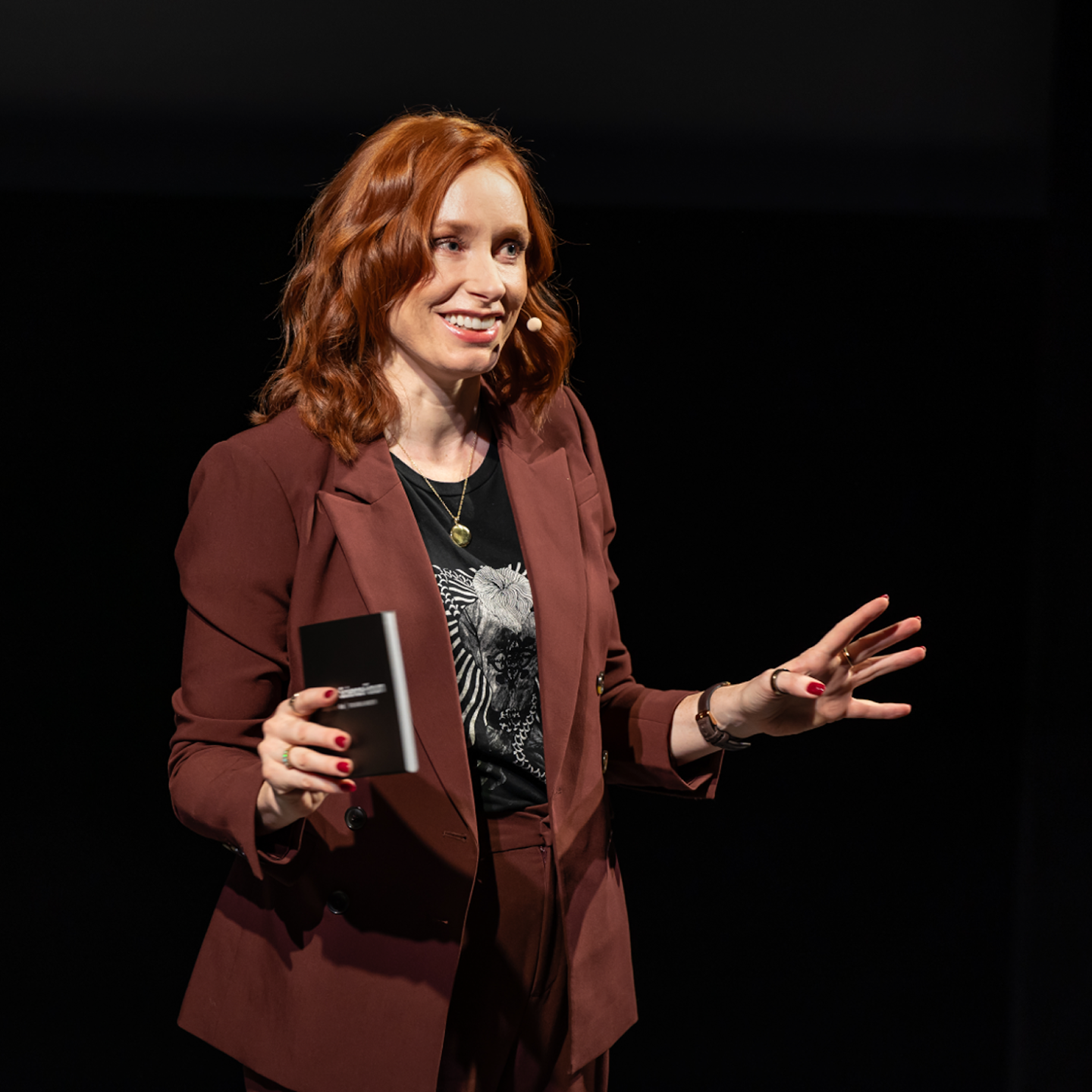 A medium shot of Hannah Fry, a woman with wavy, shoulder-length red hair, speaking. She is smiling and wearing a dark reddish-brown suit over a black t-shirt. She holds a small black book in one hand and gestures with the other against a black background.