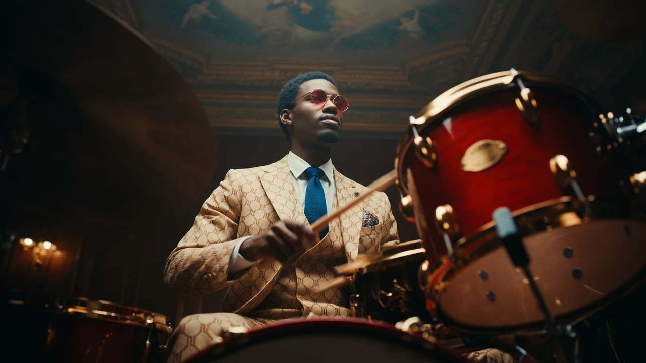 A stylish man in a patterned suit and red sunglasses playing the drums.