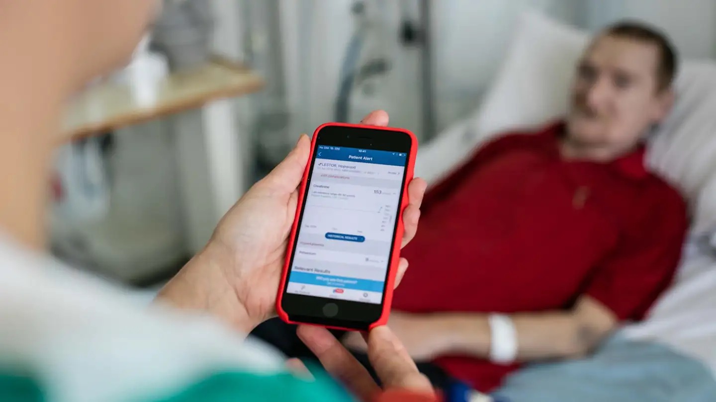 Medical professional stands by a patient in a bed. The medical professional is using the Streams app to monitor the patient.