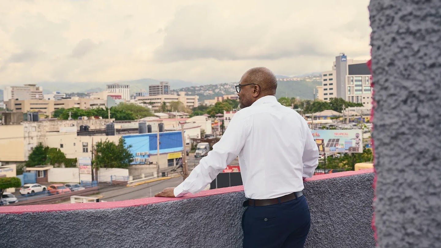 Evan Thompson, Principal Director of the Meteorological Service Jamaica, looking out over the city of Kingston under an overcast sky from a rooftop balcony.