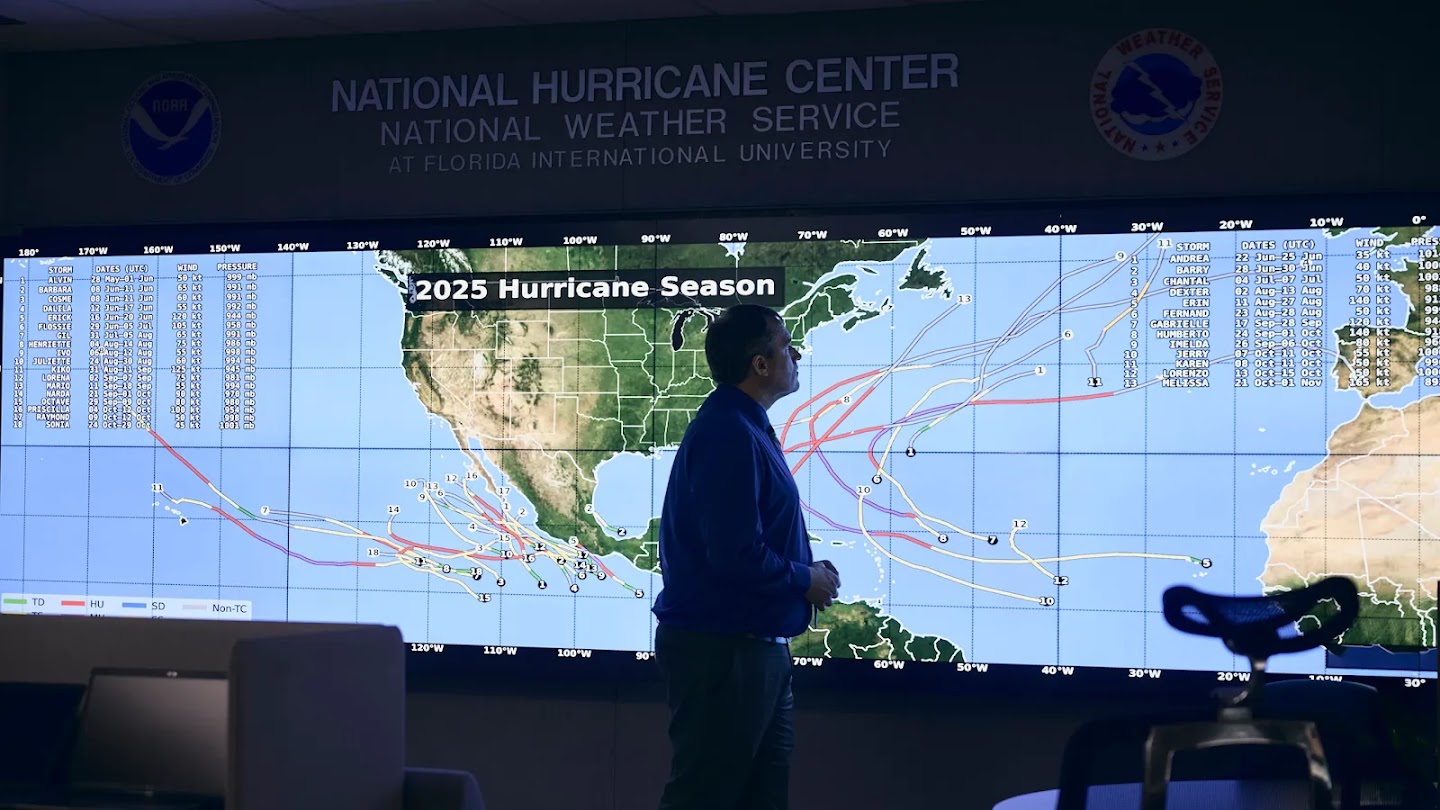 A man in a blue button-down shirt stands in front of a large, wall-mounted digital map titled "2025 Hurricane Season" at the National Hurricane Center. The map displays numerous red and pink lines tracking the paths of various hurricanes across the Atlantic Ocean, with labels for storms such as "MELISSA," "HUMBERTO," and "GABRIELLE" visible on the screen.