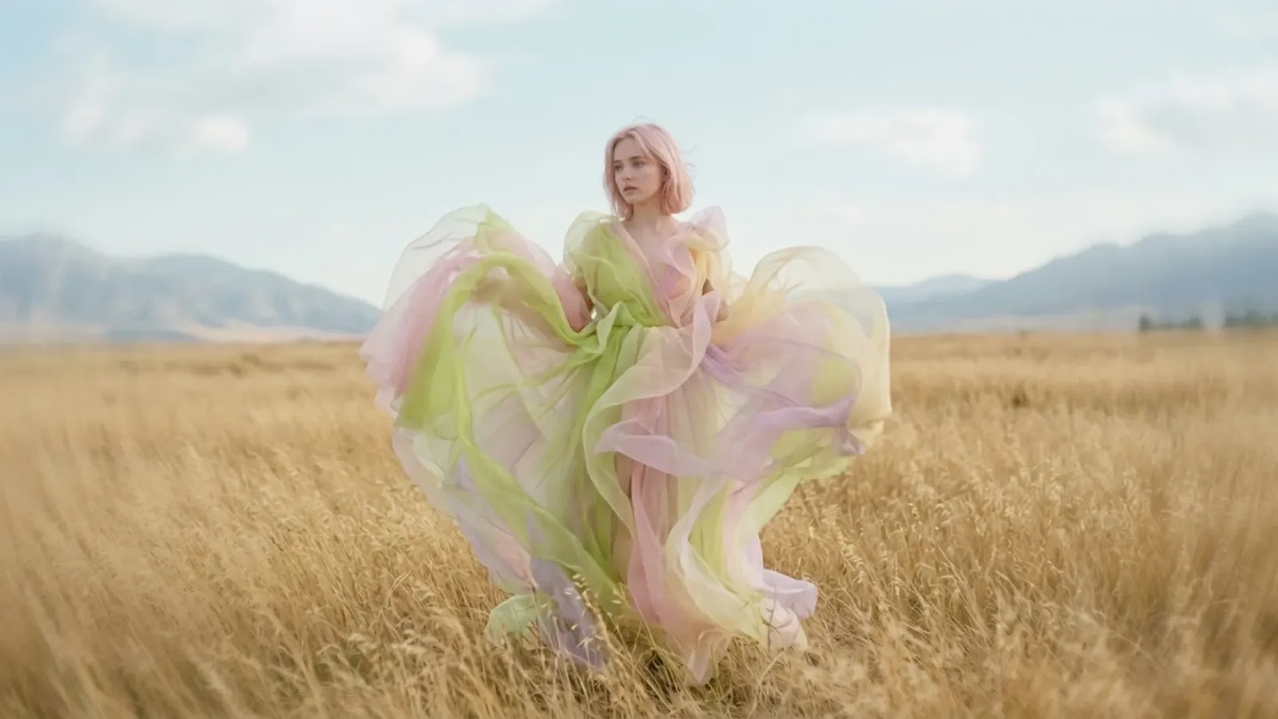 A young woman with short pink hair stands in a vast, golden wheat field under a pale blue sky. She wears an ethereal, voluminous gown made of translucent layers of pastel green, pink, and yellow fabric that billow out around her in the wind.