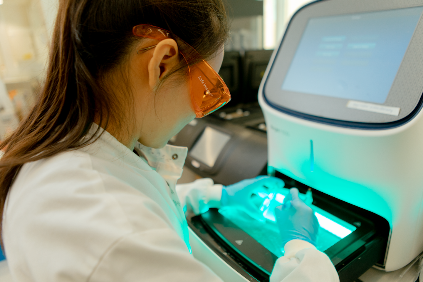 A scientist wearing goggles examines an object illuminated from underneath, by a harsh blue-tinted light.