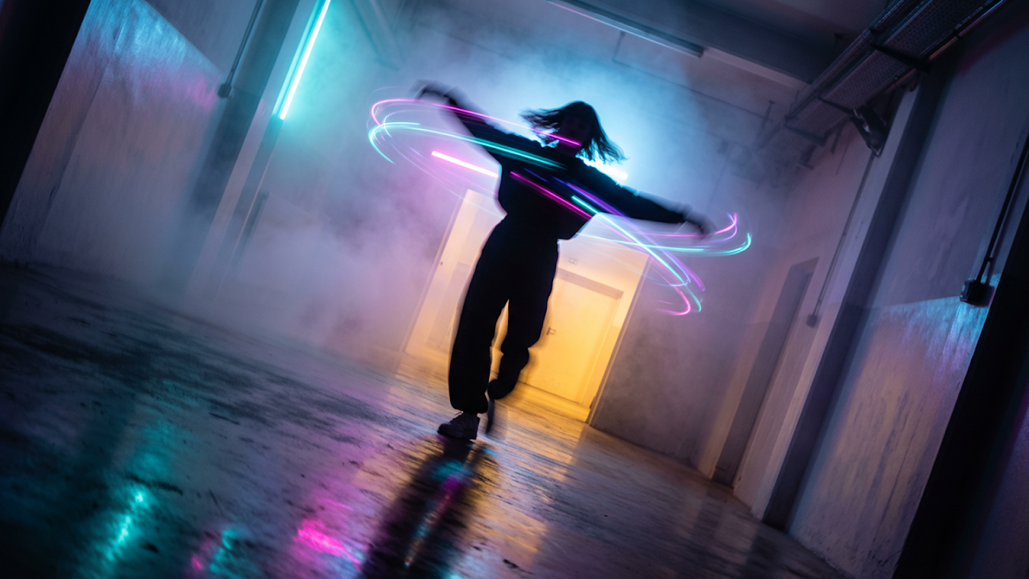 Long-exposure photography of a dancer in a dark industrial corridor, surrounded by swirling trails of neon pink and blue light created by their movement.