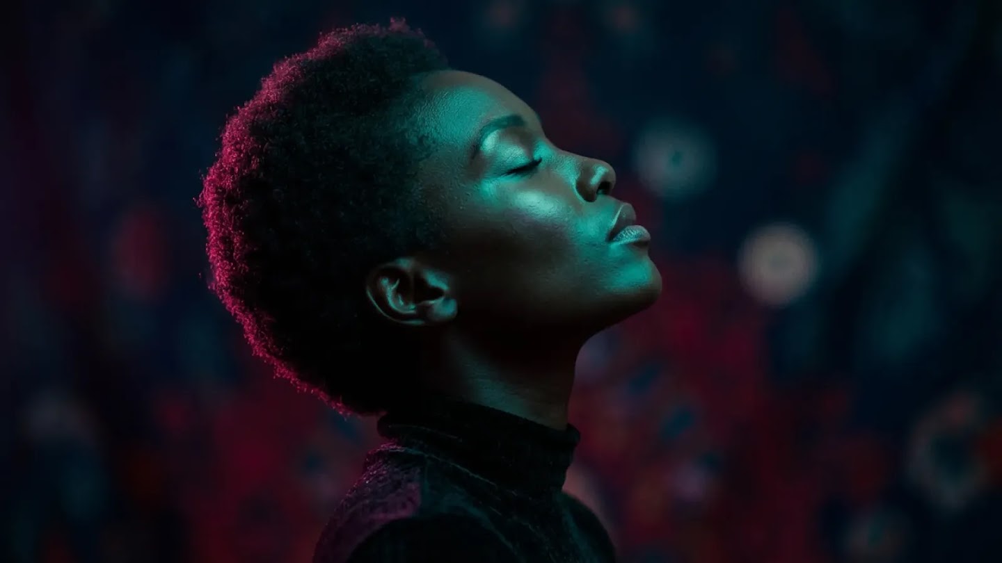 A profile portrait of a woman with short hair, illuminated by dramatic teal and magenta rim lighting against a dark, textured background. Her eyes are closed, and the high-contrast lighting emphasizes the contours of her face and the silhouette of her head.