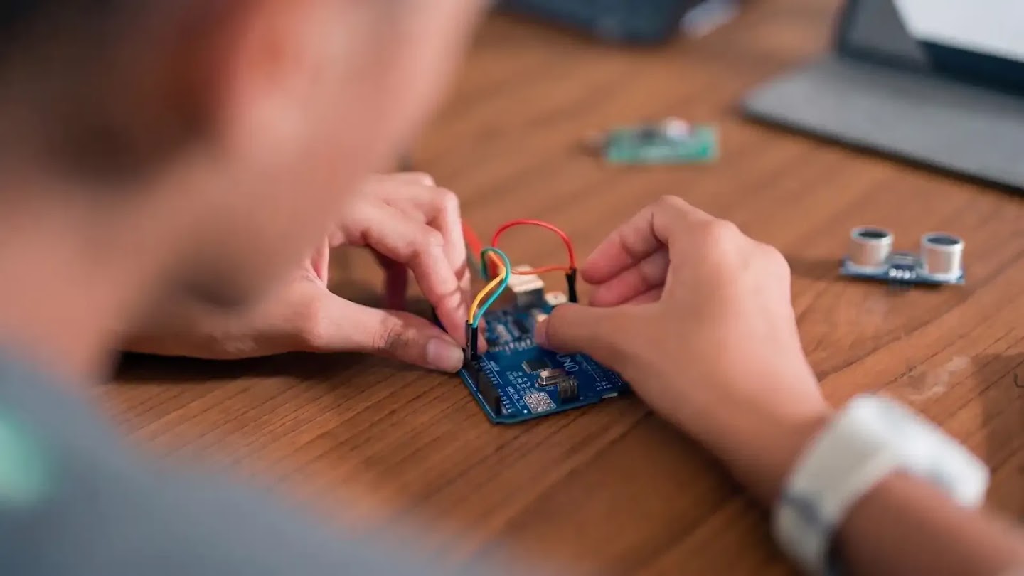 Close-up of a person's hands connecting colored jumper wires to an Arduino Uno microcontroller board on a wooden table.