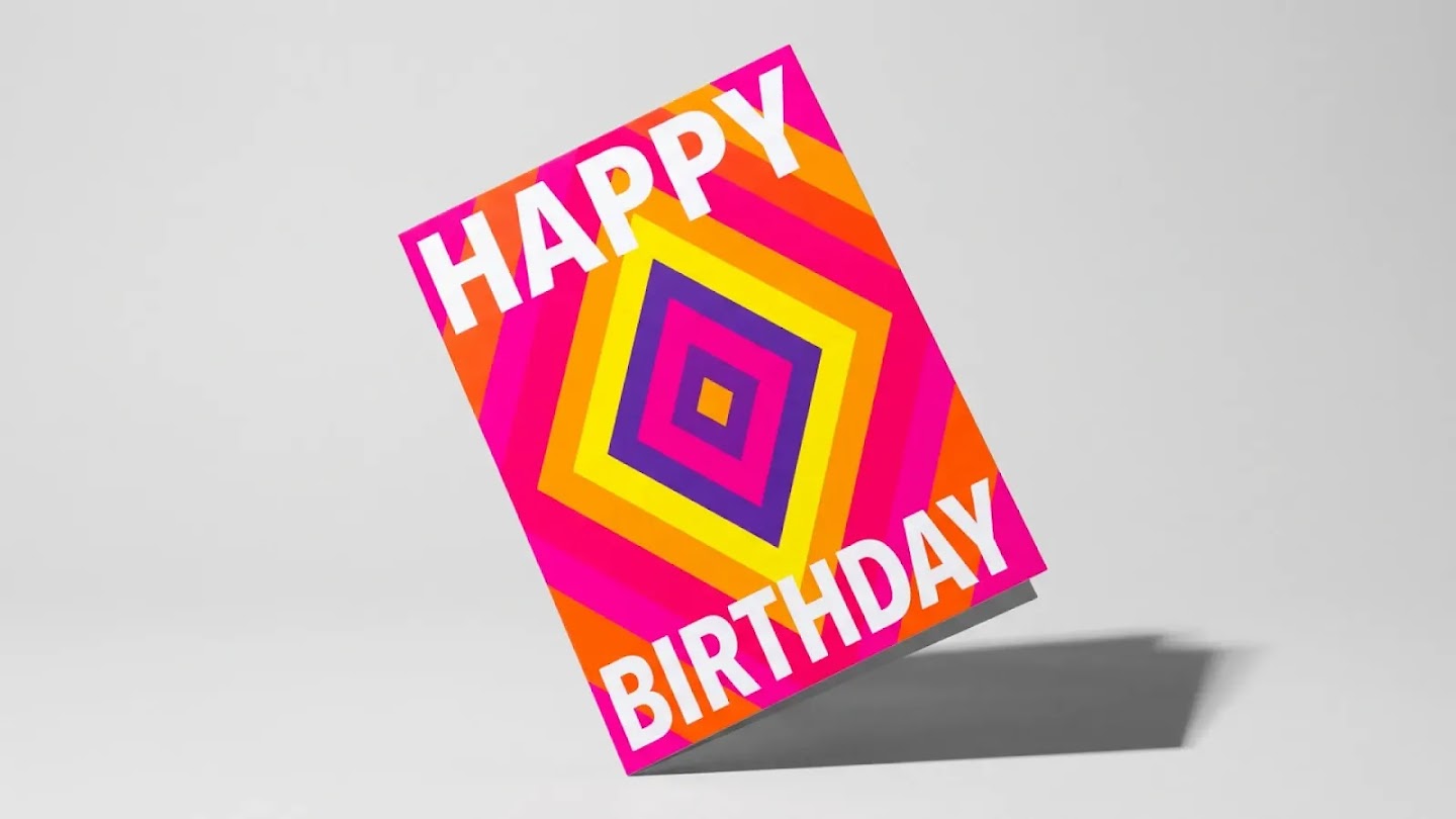 A vibrant pink birthday card is shown at a slight angle. The center features a series of concentric diamond shapes in orange, yellow, and purple. The words "HAPPY" and "BIRTHDAY" are printed in bold, white block letters at the top and bottom.