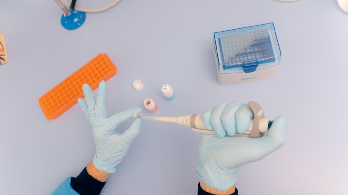 A pair of medical-gloved hands holding a pipette and receptacle.