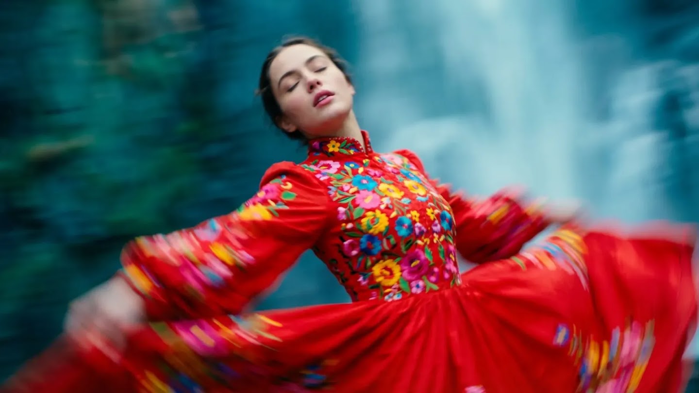 A dynamic, motion-blurred shot of a woman in a vibrant red traditional dress featuring colorful floral embroidery. She is spinning, causing the fabric of her skirt and sleeves to flare out in a circular motion. Her eyes are closed with a serene expression, set against a soft, out-of-focus background of cool blue and green tones that suggest a natural outdoor setting.