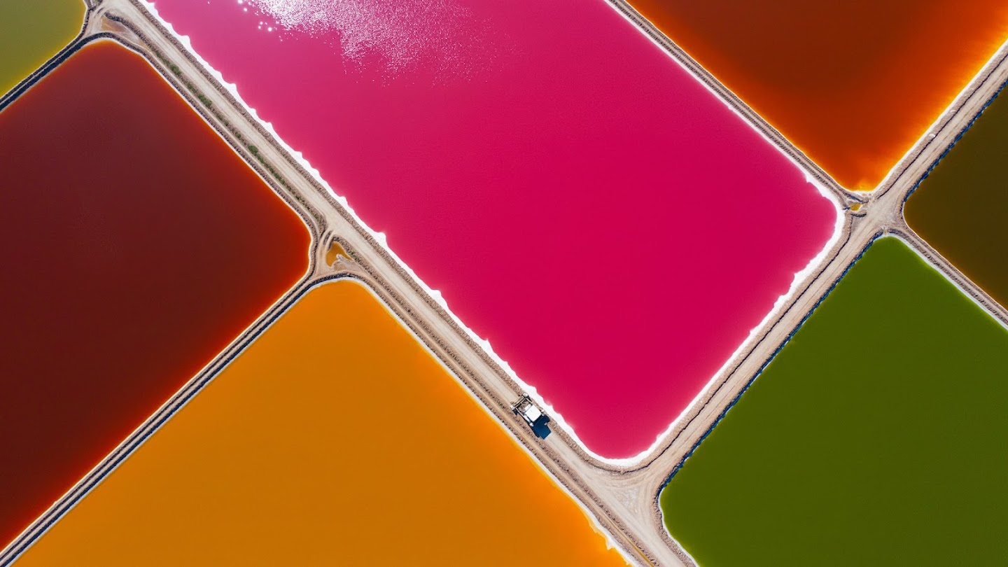 AI-generated image of: an aerial, top-down view of vibrant salt evaporation ponds arranged in a geometric grid, featuring intense, contrasting colors like vivid magenta, deep rust, mustard yellow, and lime green, separated by thin, textured earthen ridges