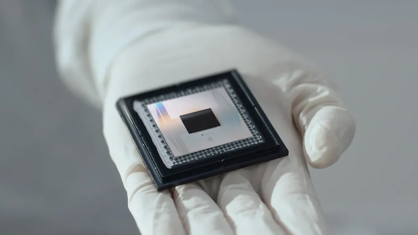 A gloved hand holding a square microchip processor with a reflective surface in a cleanroom setting.