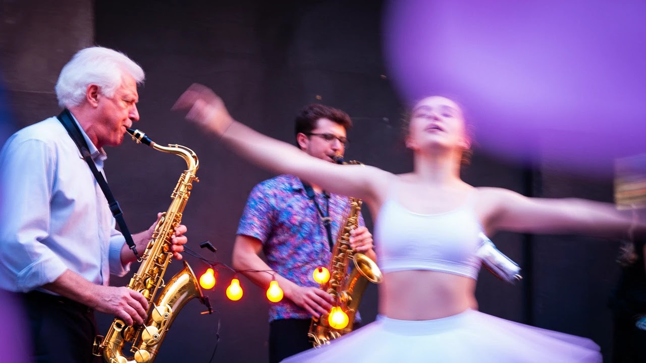 A ballerina spins in front of two saxophone players during a performance.