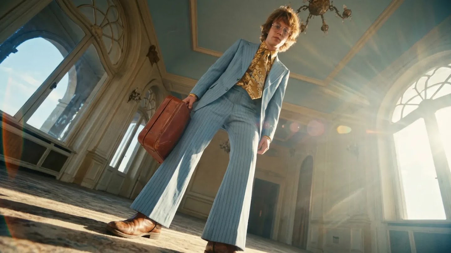 A low-angle, wide-lens shot of a person in a vintage-style blue pinstripe suit and a patterned gold tie. They are standing in a grand, sun-drenched room with high ceilings and ornate windows, holding a brown leather suitcase as light streaks across the frame.