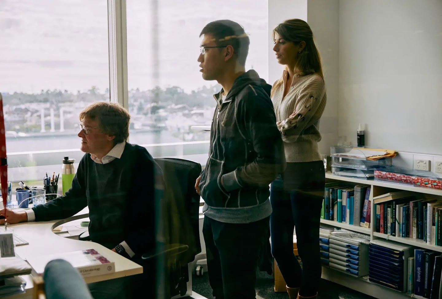 A photograph of a people inside David Komander's office. We are looking through the glass from the corridor. Two people are standing behind Komander and watching what he is doing on the computer.
