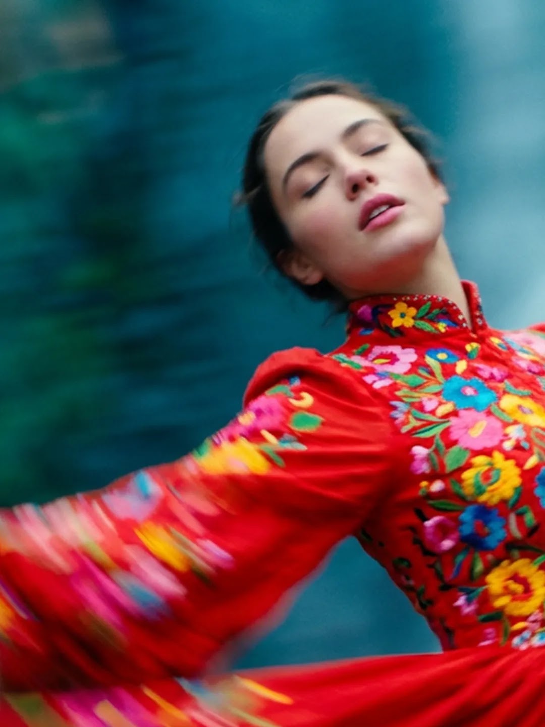 A dynamic, motion-blurred shot of a woman in a vibrant red traditional dress featuring colorful floral embroidery. She is spinning, causing the fabric of her skirt and sleeves to flare out in a circular motion. Her eyes are closed with a serene expression, set against a soft, out-of-focus background of cool blue and green tones that suggest a natural outdoor setting.