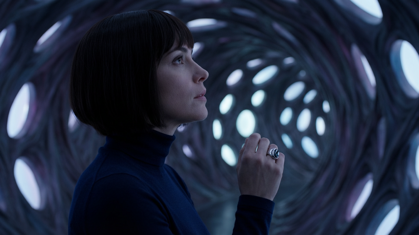 Cinematic close-up profile of a woman with short dark hair standing inside a futuristic, organic structure with glowing, cellular-patterned walls.