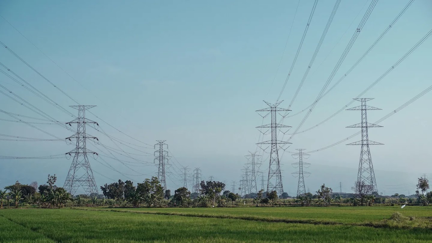 A wide landscape shot of high-voltage power lines and transmission towers stretching across a lush green field under a clear blue sky.
