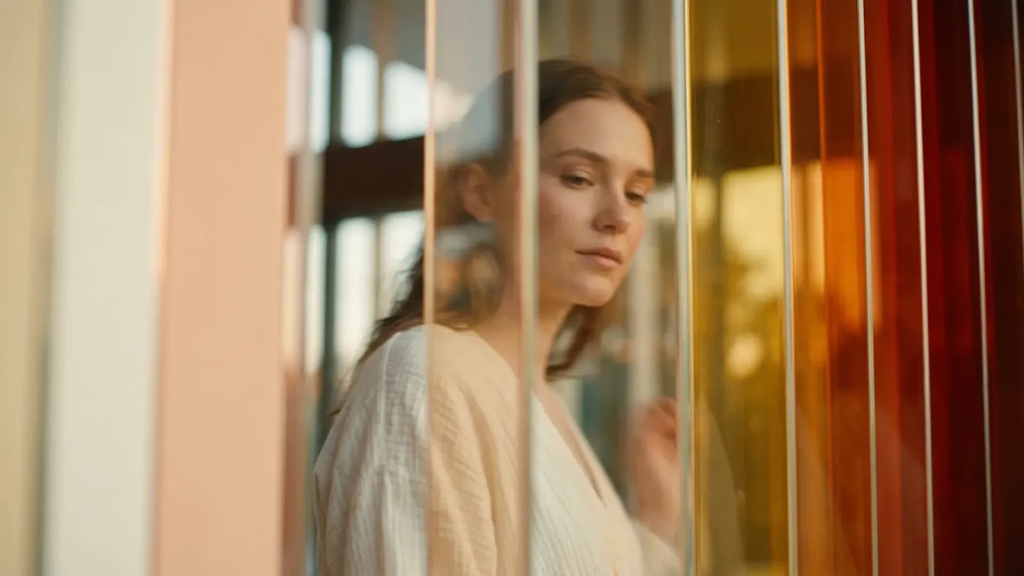 A soft-focus portrait of a woman viewed through a series of vertical glass panels tinted in shades of orange, yellow, and clear. She has a contemplative expression and wears a light-colored, textured garment, creating a layered and atmospheric effect.