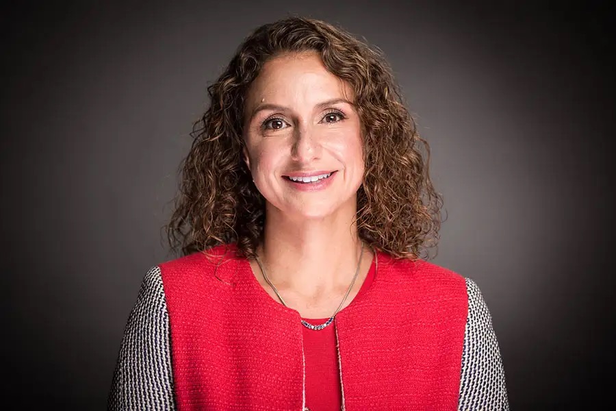 Lila Ibrahim dressed smartly and smiling in front of a plain, dark grey background.